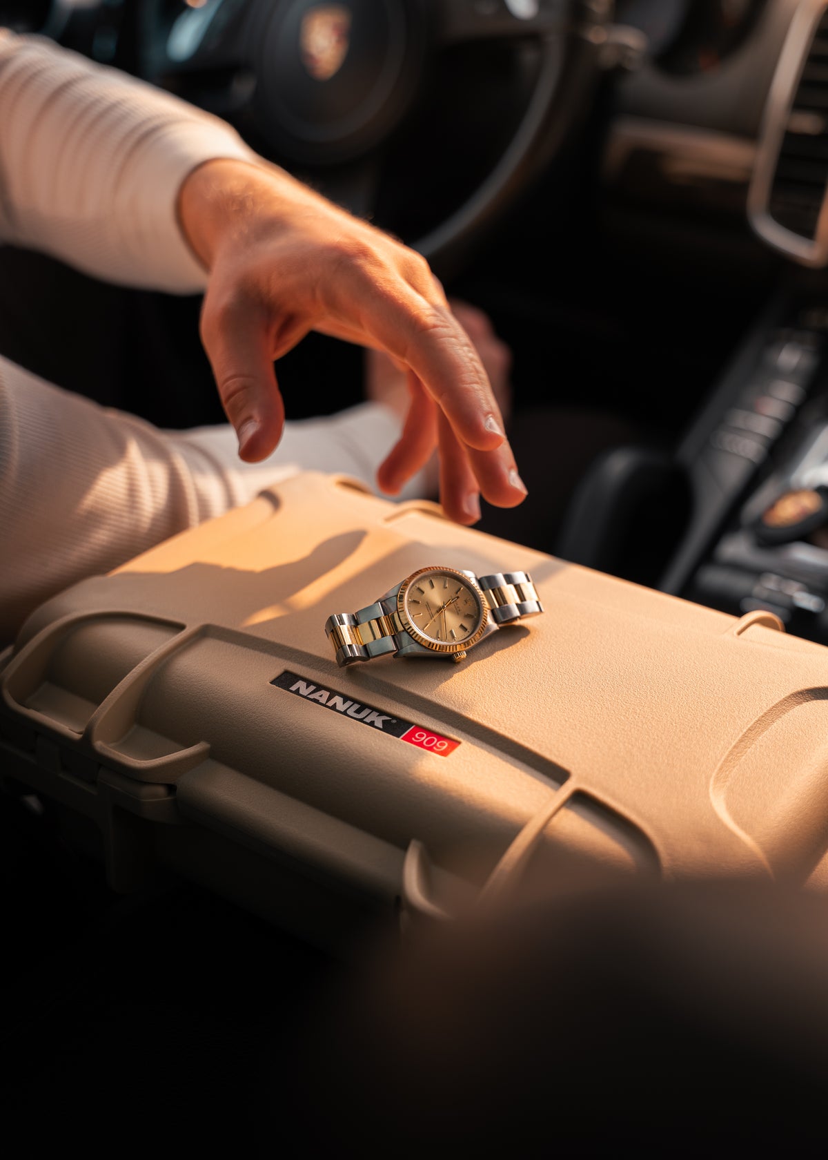 A silver-toned metal watch with a gold dial rests on the lid of a beige Naniux hard case, with a person's hand reaching toward it inside a car.