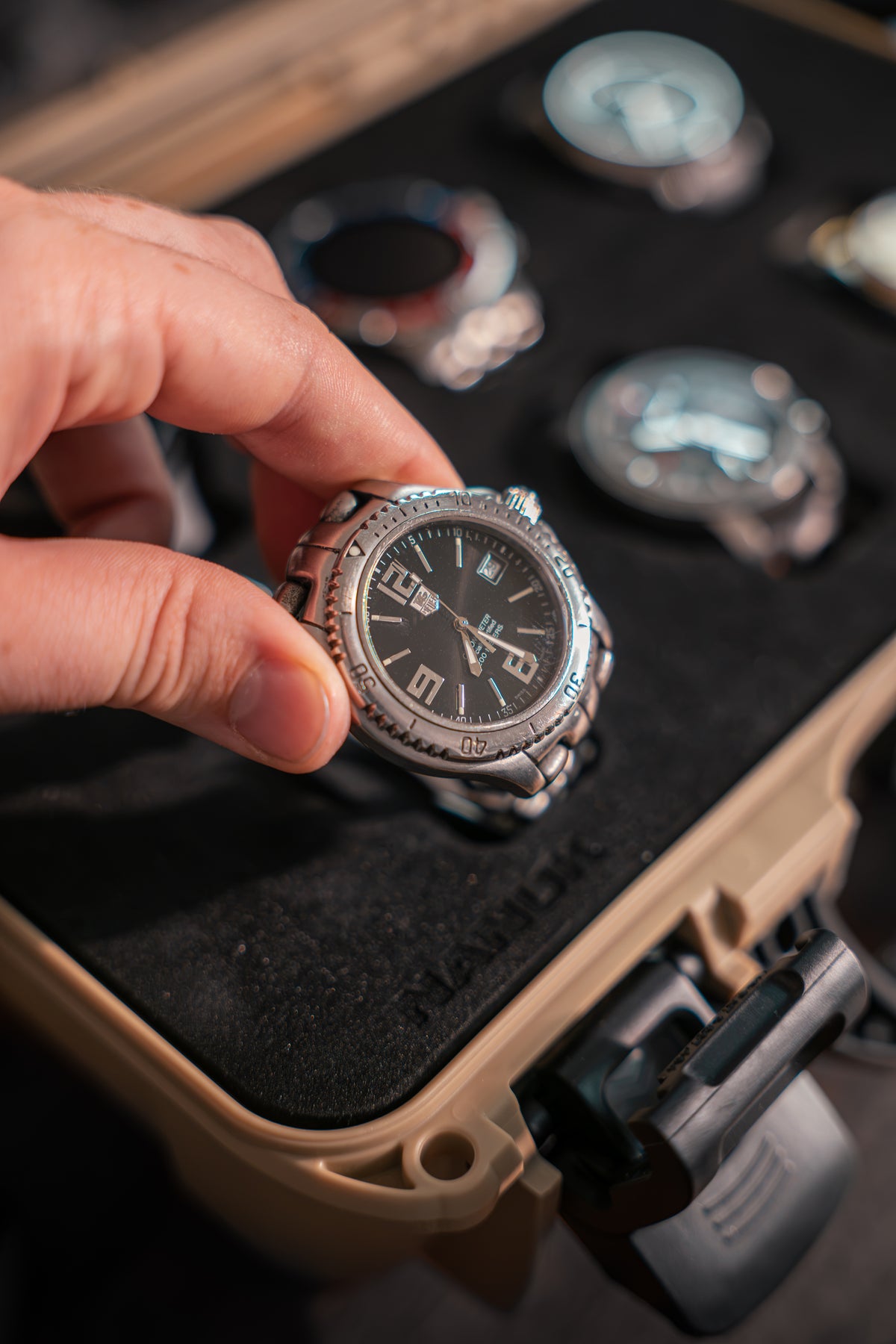 A hand holding a silver stainless steel wristwatch with a black dial and bezel, positioned over a tan hard case with other watches inside.