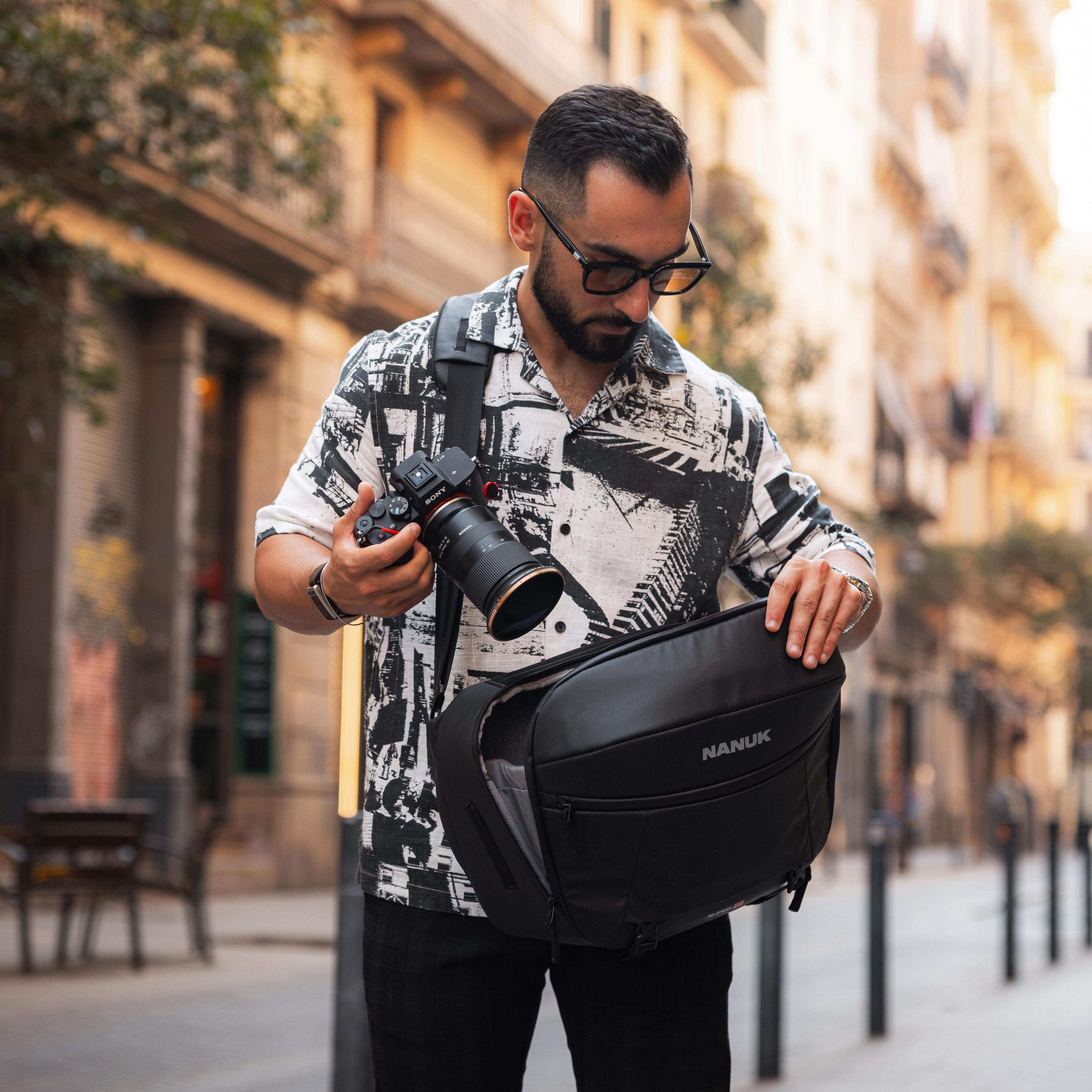A man wearing a black and white patterned shirt and glasses carries a black Nanuk camera bag while holding a DSLR camera on a city street.