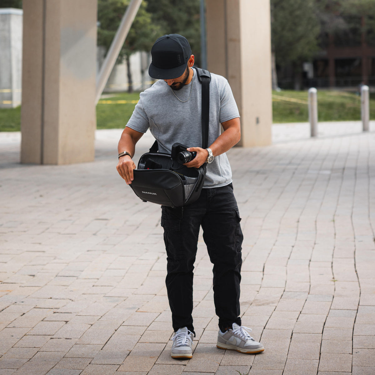 A man wearing a black cap, gray t-shirt, and black cargo pants is shown outdoors on a paved walkway, holding a black camera bag with a DSLR camera inside. The bag has a white logo that reads 'BAGS'.