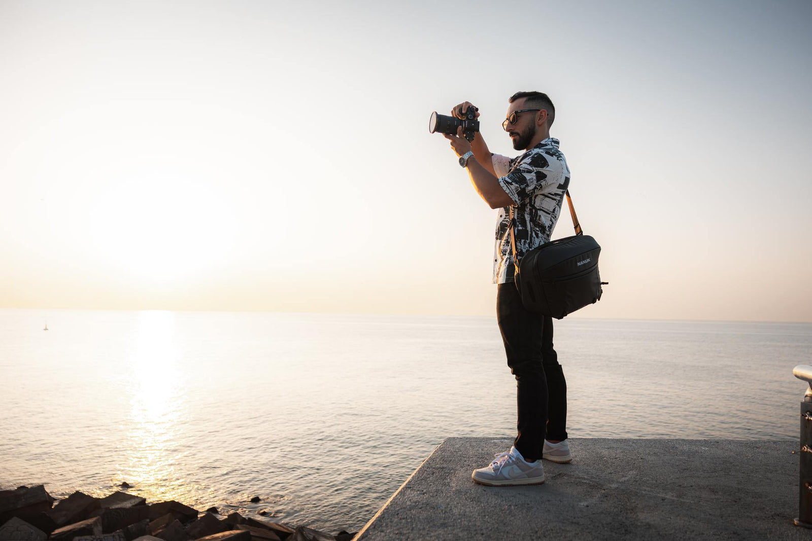 A man wearing a black and white patterned shirt, black pants, and white sneakers stands on a concrete pier overlooking the ocean at sunset, holding a camera and carrying a black shoulder bag.