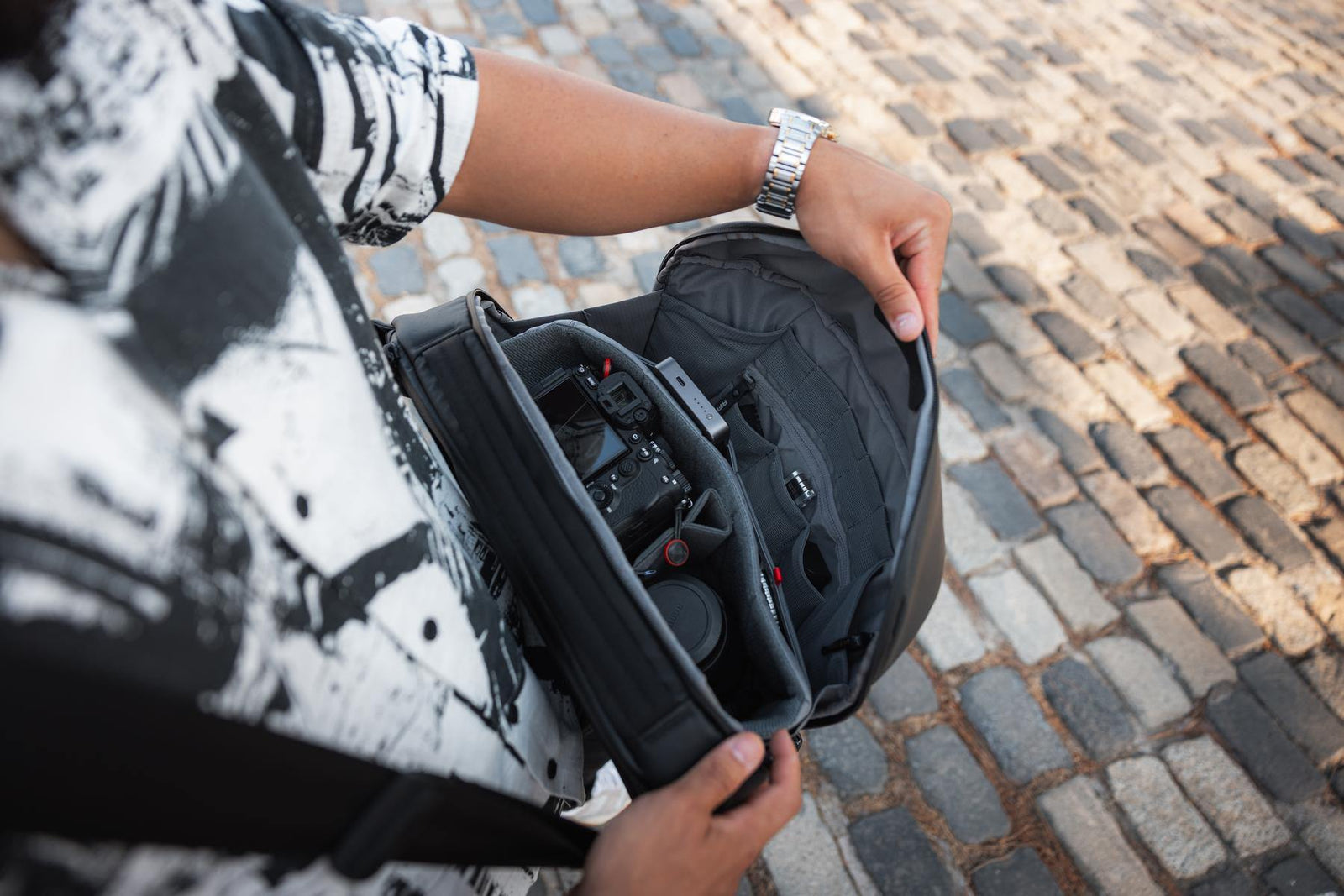 A close-up of a person holding open a black camera backpack or gear bag, showing the interior organizer and a camera inside, with a cobblestone street in the background.