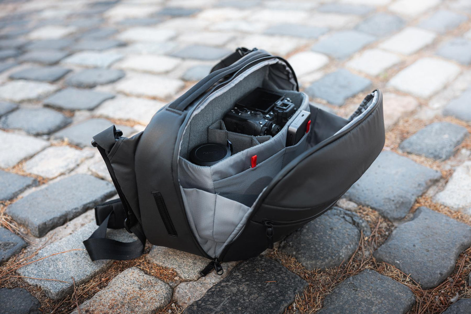 A black and gray camera bag lies open on a cobblestone surface, with a camera and lens visible inside the padded compartment.
