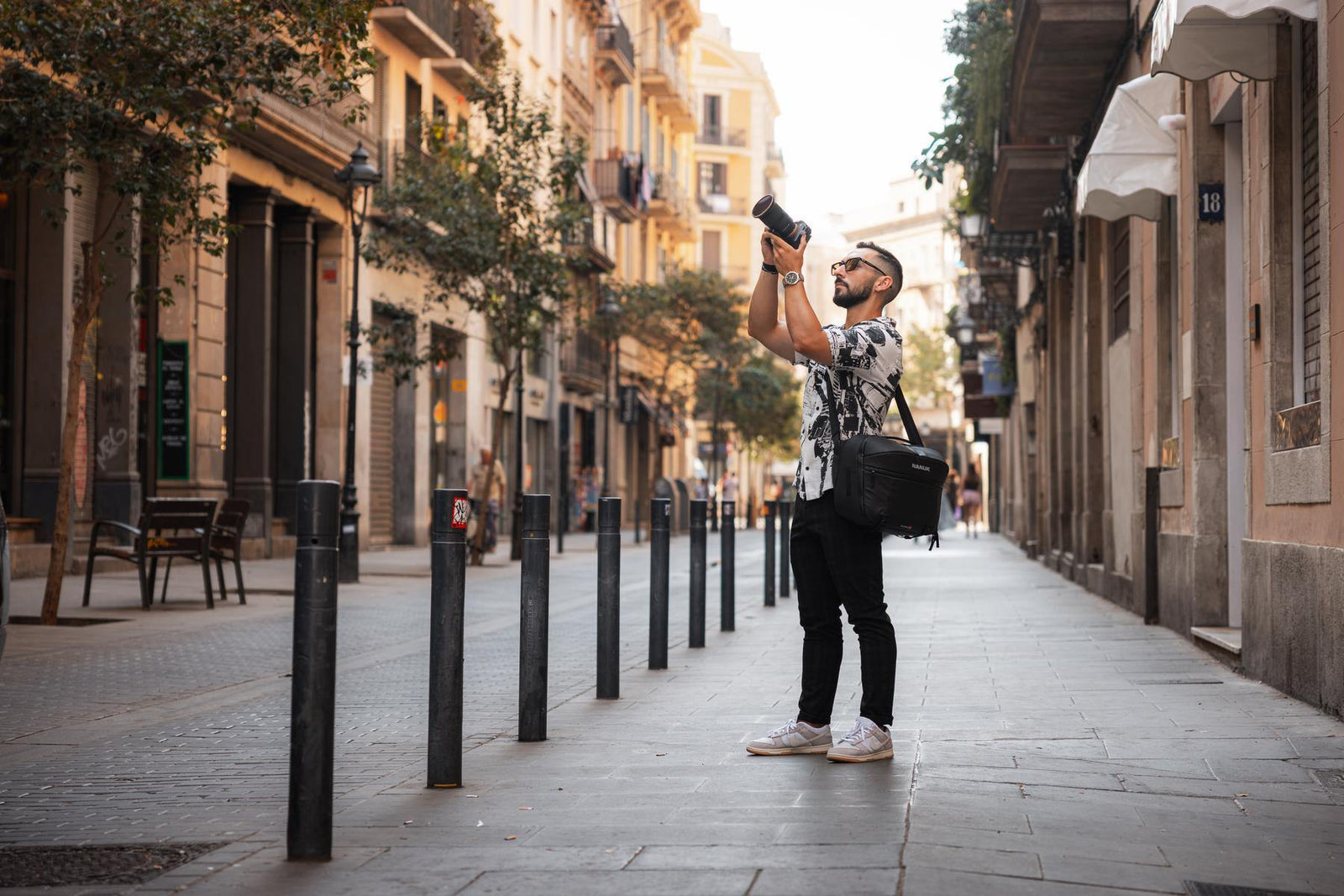 A man wearing a black and white patterned shirt, black pants, and white sneakers stands on a city street taking a photo with a camera. He has a black shoulder bag slung over his shoulder.