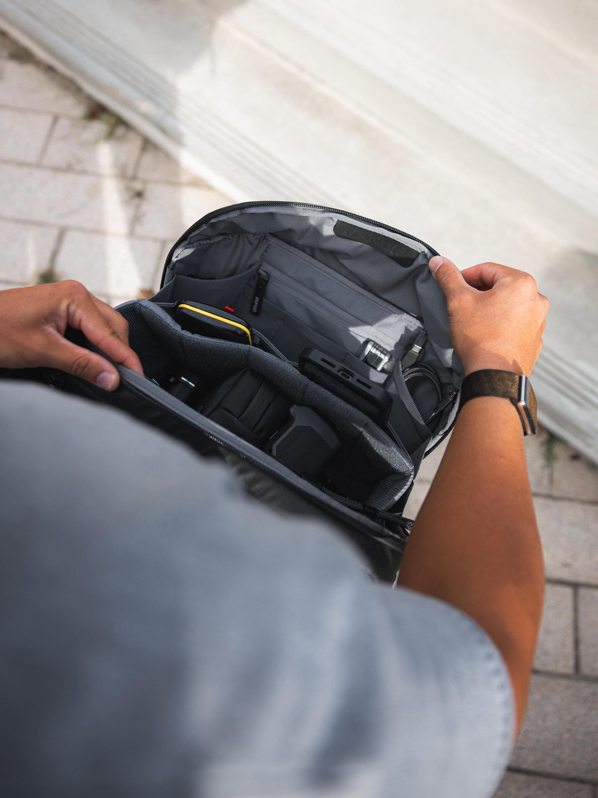 A close-up of a person's hands opening a dark gray camera backpack, showing the interior organization with padded compartments and pockets. The person is wearing a light blue-gray t-shirt and a black watch, standing outdoors on a paved surface next to a white wall.