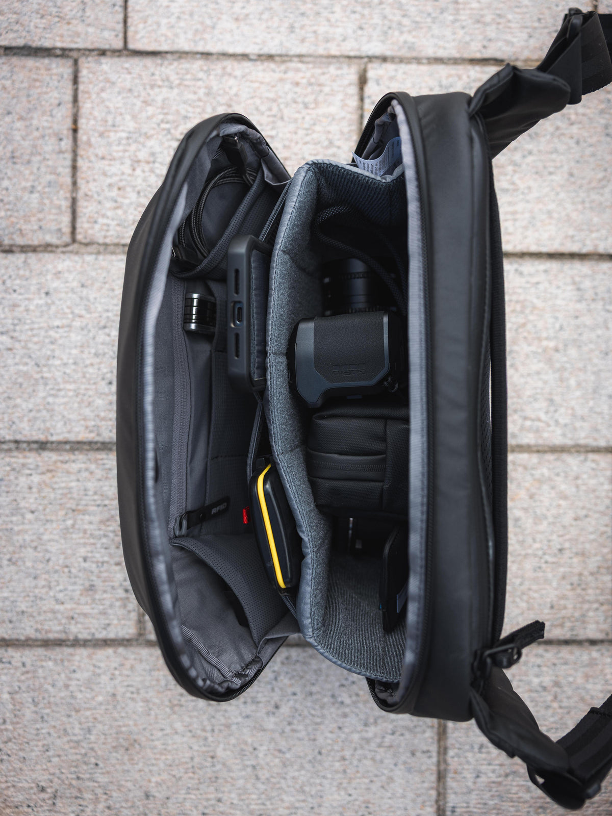 An open black camera bag laid flat on a light gray stone surface, showing padded interior compartments and a camera accessory inside.