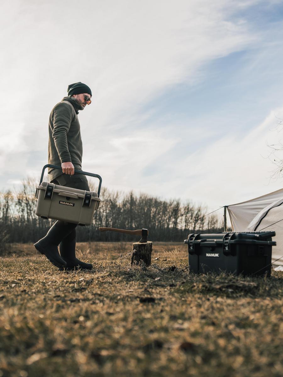 Man carrying a cooler box in an outdoor setting with a tent and fireplace.