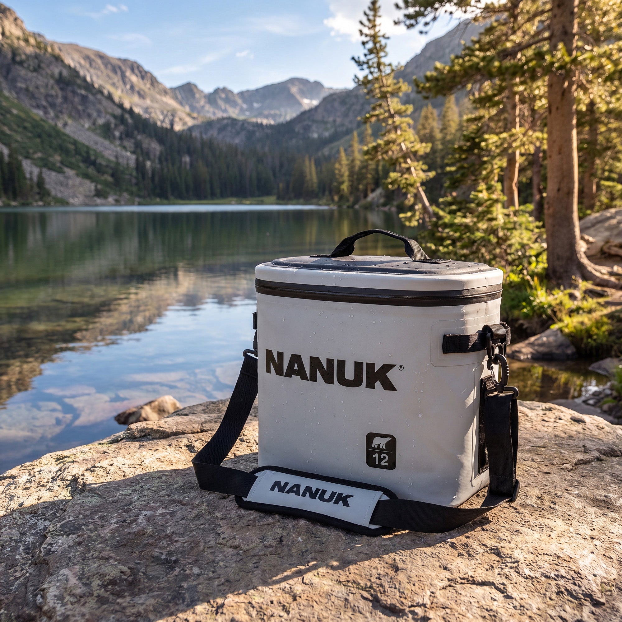 A white and black Nanuk 12-inch cooler bag sits on a large rock in a mountain lake setting, with pine trees and rugged peaks in the background.
