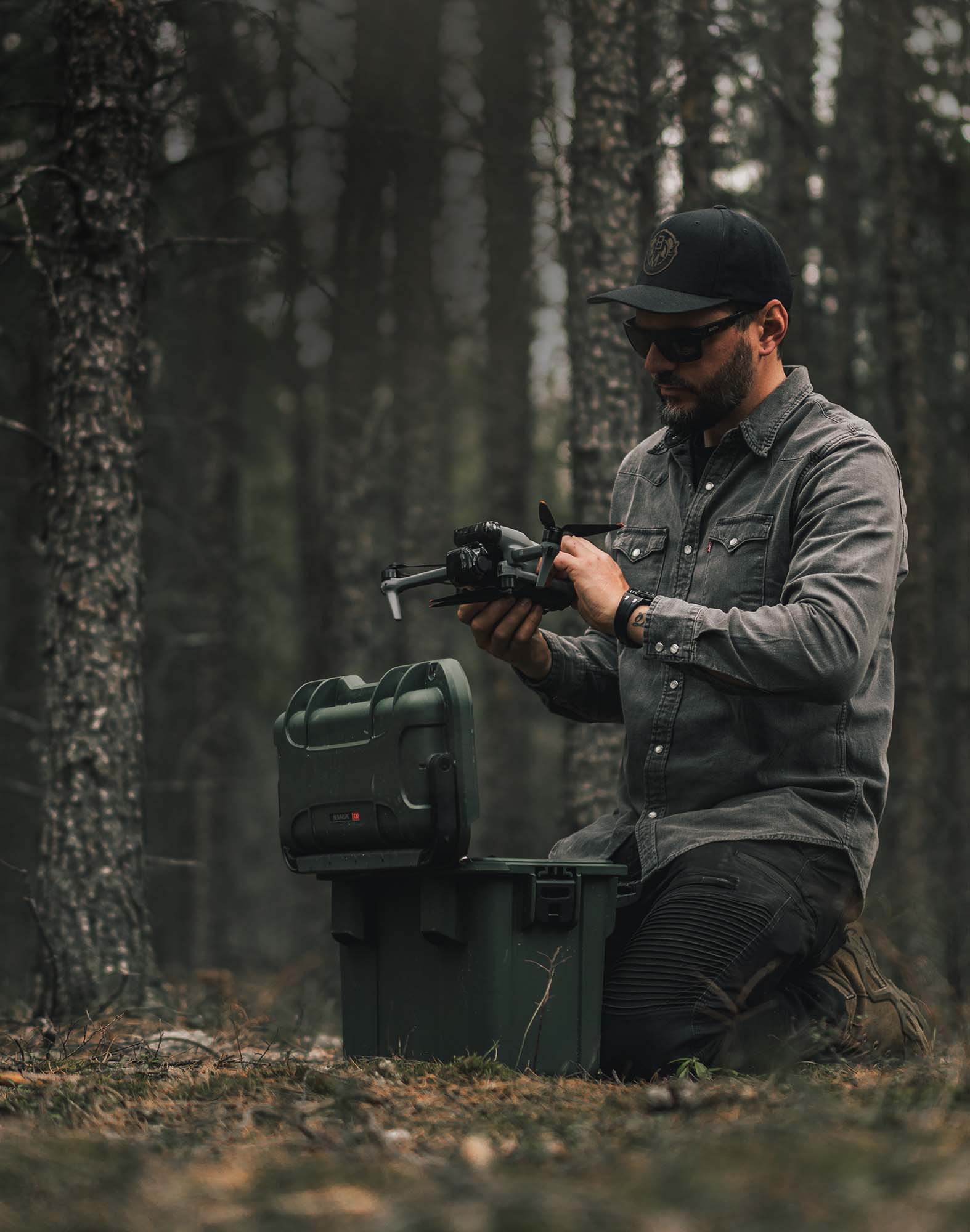 Man kneeling in the woods preparing a drone next to an olive green NANUK protective hard case.