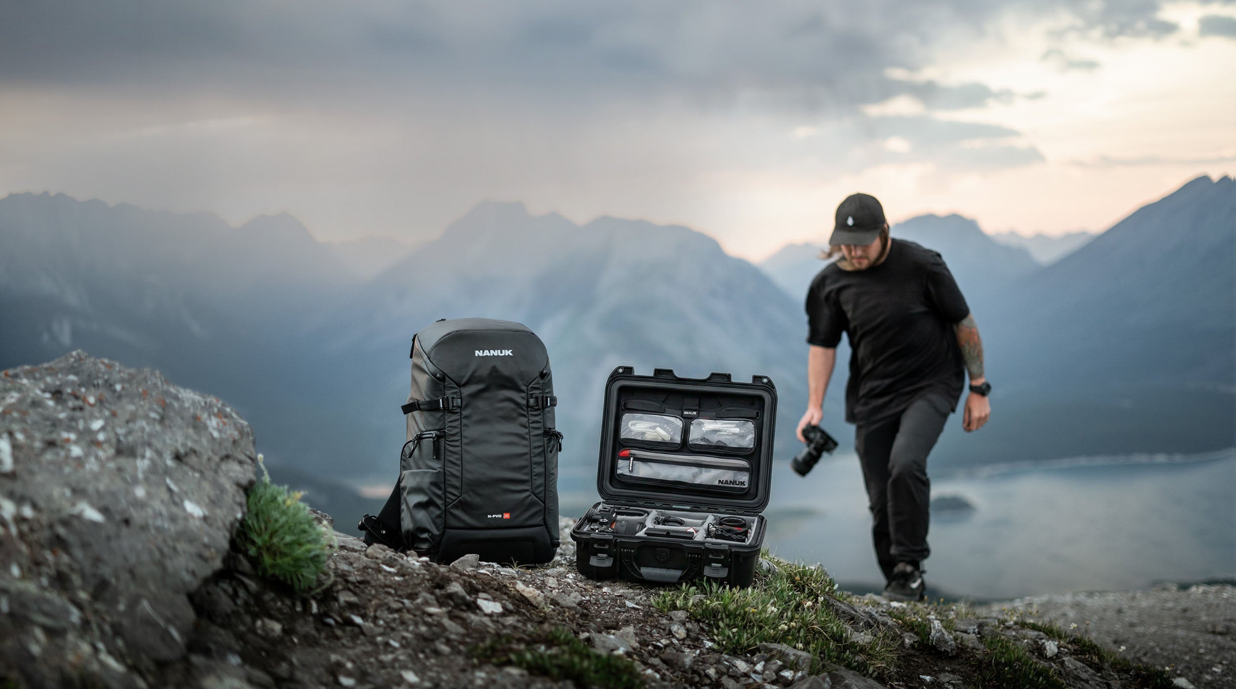 A NANUK camera backpack and an open hard-shell protective case with photography gear on a rocky mountain ridge with a photographer walking in the background.