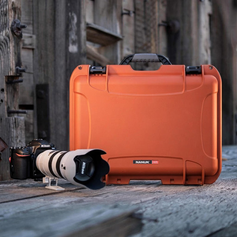 An orange hard camera case with a black handle and latches sits on a weathered wooden surface, with a DSLR camera and lens placed in front of it. The case has a visible NARUK logo.