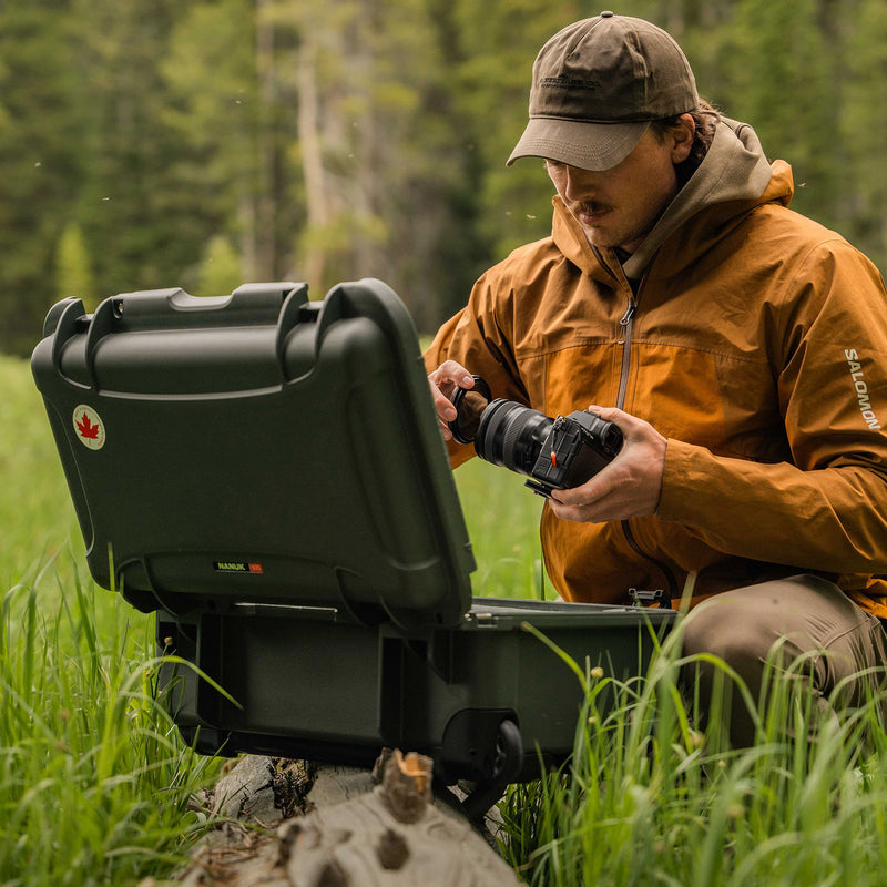 A man in an outdoor forest setting wearing an orange hooded jacket and a brown cap, kneeling beside an open black hard case and holding a camera. The jacket has a visible 'SOLIS' brand mark on the sleeve.