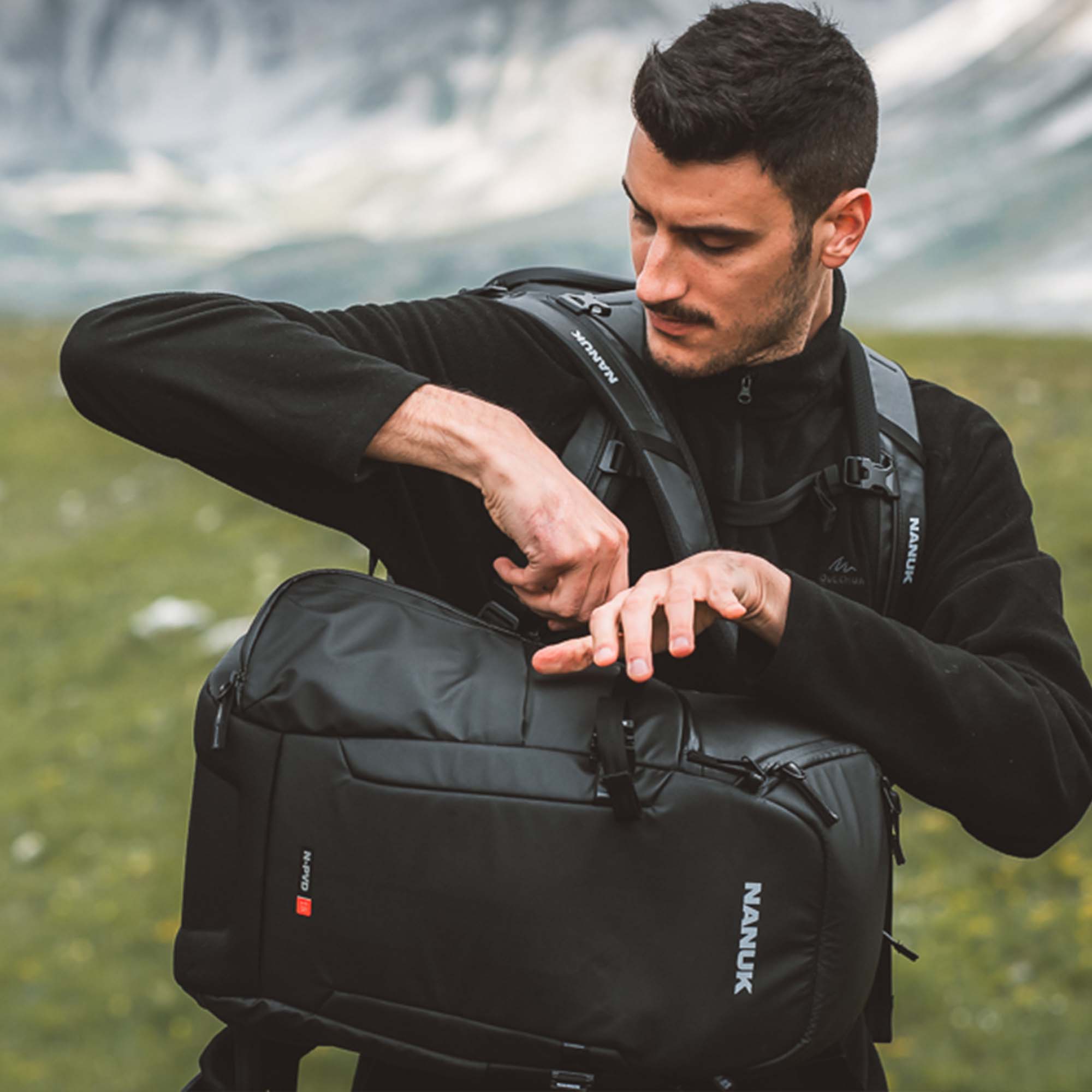 Man adjusting a black backpack outdoors with mountains in the background