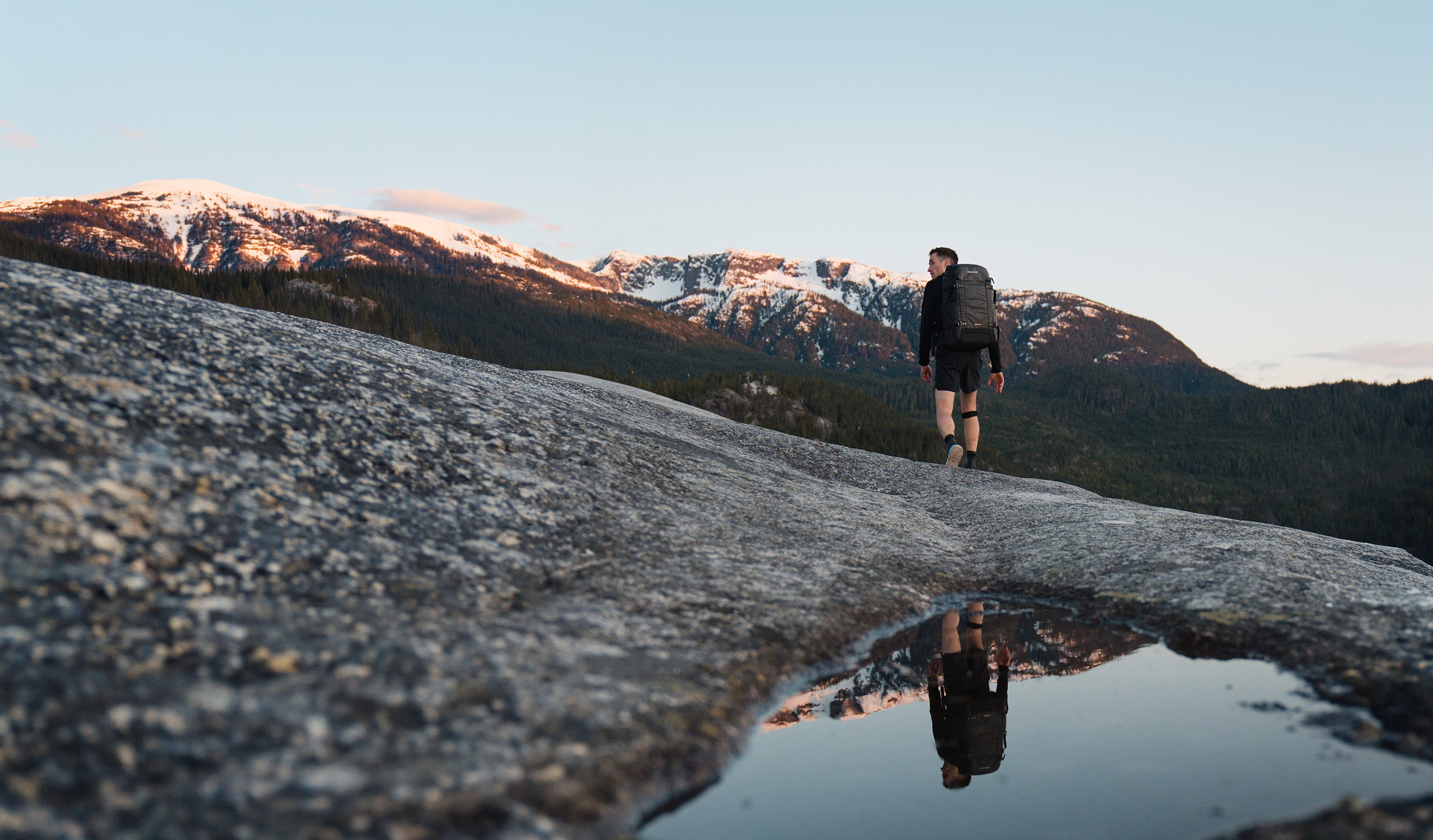 A hiker with a large black backpack walking on a rocky mountain summit, reflected in a small puddle with snow-capped peaks in the distance.