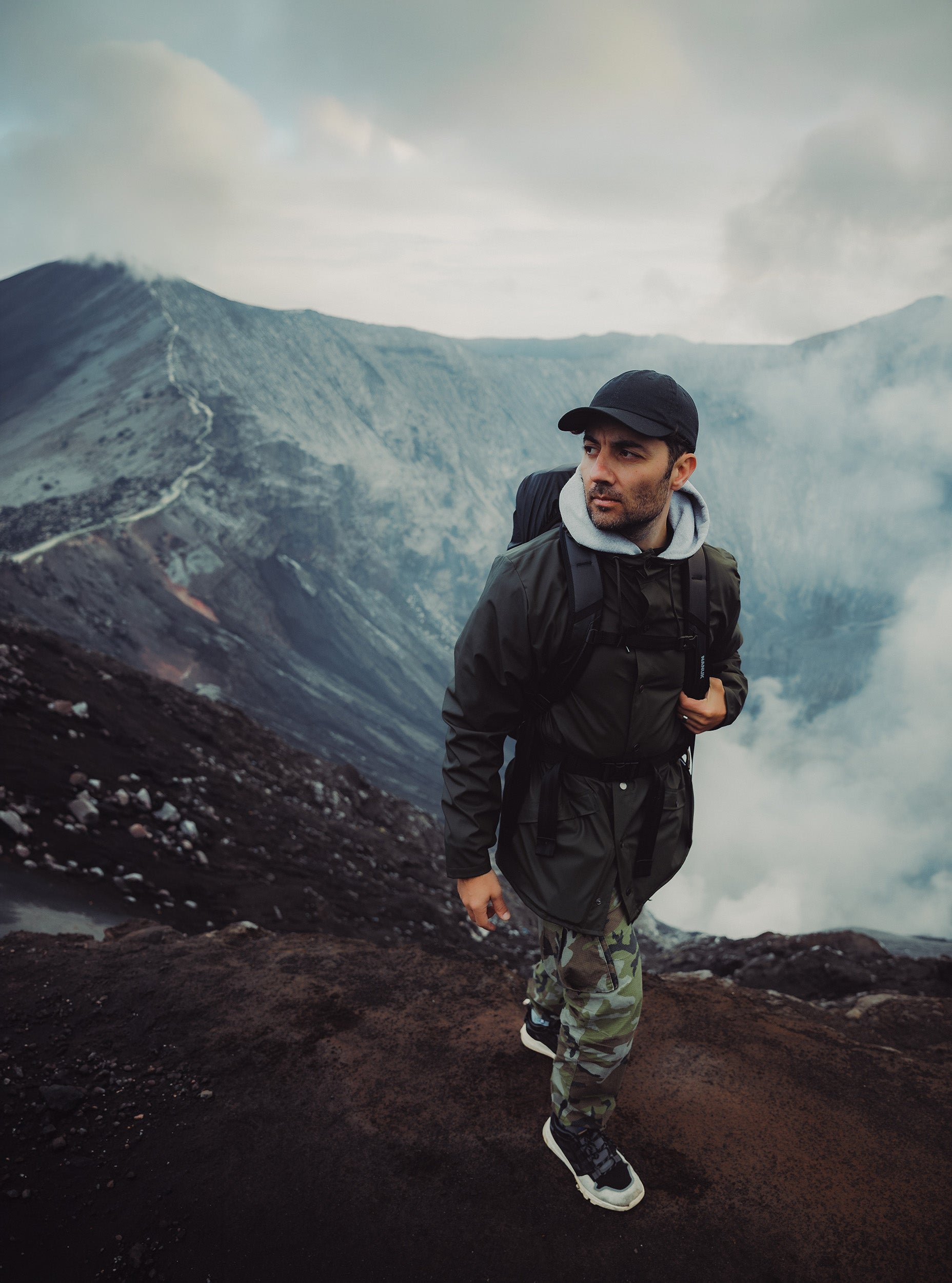 A man in a dark green jacket and camouflage pants hikes along the rocky rim of a volcanic crater under a misty, overcast sky.