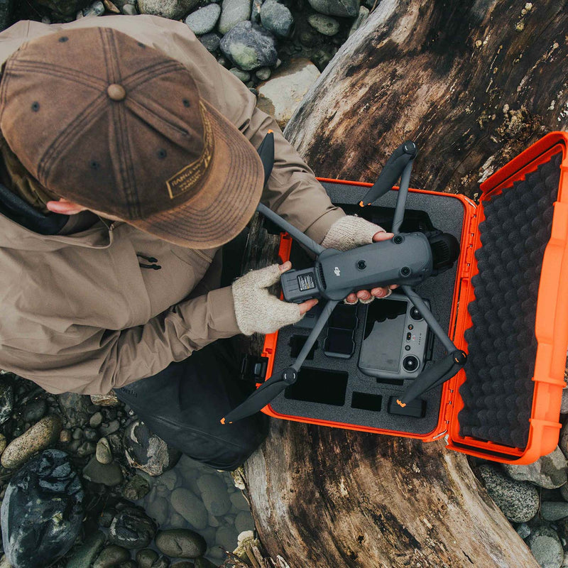 A person in a brown cap and tan jacket handles a gray drone next to an open bright orange hard case with foam lining, set on a rocky outdoor surface.