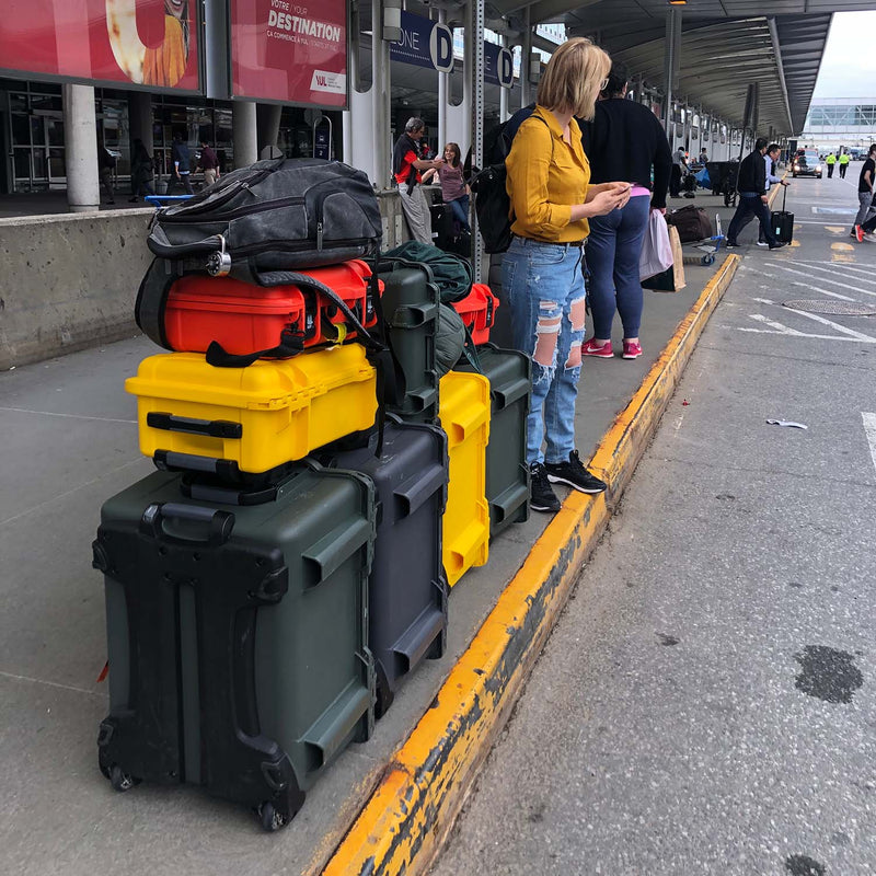 A stack of hard-shell carry-on luggage in dark green, yellow, red, and black sits on a airport departure curb next to a woman in a yellow top and ripped blue jeans looking at her phone. Other travelers and airport terminals are visible in the background.