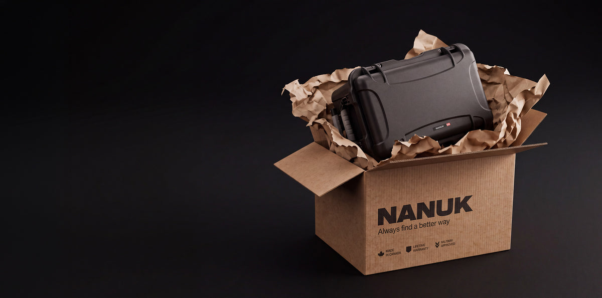 A black Nanuk protective case nestled inside a brown cardboard box lined with crumpled brown paper, set against a solid black background. The brand name 'NANUK' is clearly printed on the front of the box.