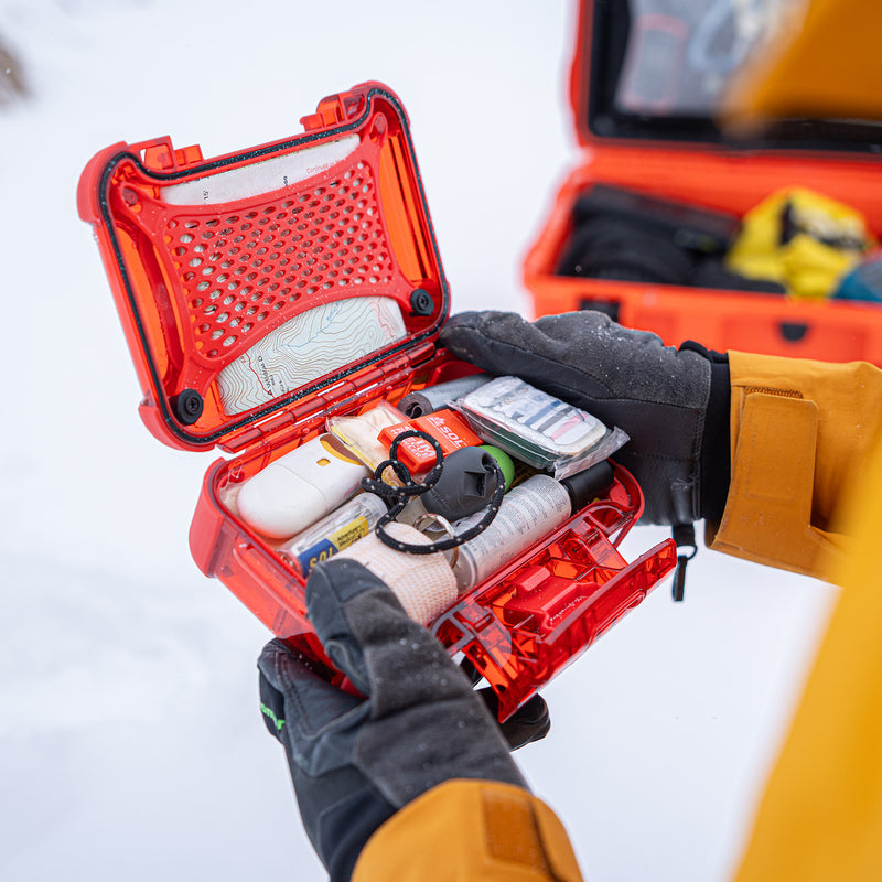 A person wearing black gloves holds an open bright red hard case filled with emergency or medical supplies, including a tourniquet, gauze, and other packaged items. Another red case sits blurred in the background on a snowy white surface.