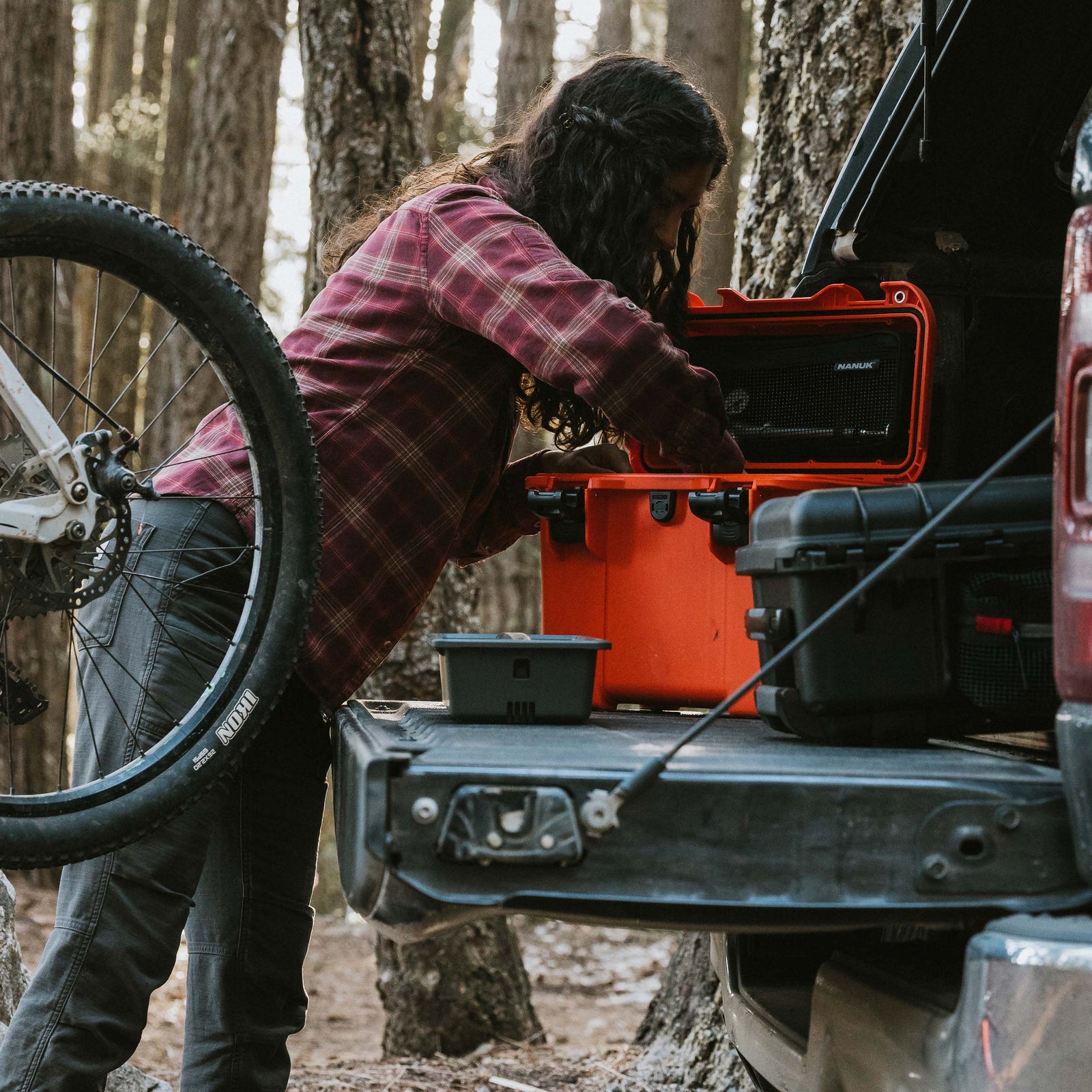 Cyclist loading a bright orange NANUK Trail 15 case into a pickup truck bed next to a mountain bike in a forest setting.