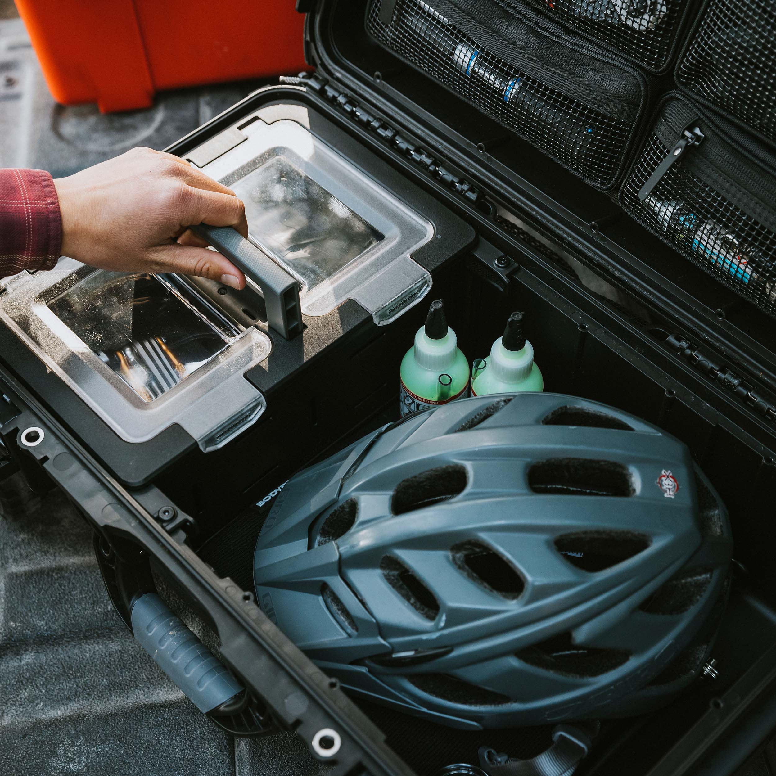 Interior of an open black NANUK Trail 30 case on a vehicle floor, showing a bike helmet, water bottles, and other gear organized beneath a clear lid.