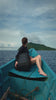 A woman wearing a dark long-sleeved top and sunglasses, with her hair in a braid, sits in the bow of a rustic blue boat on calm ocean waters. A durable black NANUK backpack rests beside her as she gazes towards a distant, lush green island under a partly cloudy sky, depicting a tranquil tropical journey or island hopping adventure.